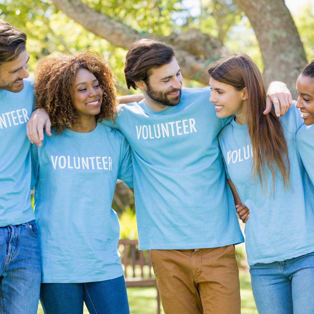 Five smiling people wearing matching blue VOLUNTEER shirts stand outdoors with arms around each other, enjoying a sunny day in a park-like setting while supporting a nonprofit for children.